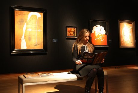 Una noia llegeix el catàleg de la subhasta de Christie's a la sala on s'exposen 'Le corps de ma brune' (1925), de Joan Miró, i ‘L'automobile fossile du Cap Creus' (1936), de Salvador Dalí. FOTO: Pous, Laura.