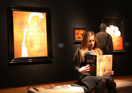 Una noia llegeix el catàleg de la subhasta de Christie's a la sala on s'exposen 'Le corps de ma brune' (1925), de Joan Miró, i ‘L'automobile fossile du Cap Creus' (1936), de Salvador Dalí. FOTO: Pous, Laura.